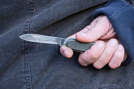 hand of a male criminal in blue black clothes and a gray green knife behind his back on the street