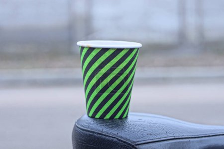 Foto de Una taza de papel rayado verde negro con soportes de café en un asiento de bicicleta de cuero sobre un fondo gris en la calle - Imagen libre de derechos