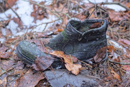 trash from one old black torn leather sneaker stands on brown dry leaves and the ground outdoors in the autumn forest