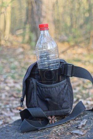 una bolsa negra hecha de tela y una botella de plástico con una bebida en el soporte de bolsillo en el suelo gris en la calle