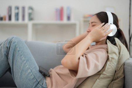 Foto de Mujer joven sonriente se relaja en un sofá con los ojos cerrados, disfrutando de su música favorita en auriculares azules en una acogedora sala de estar con una estantería en el fondo - Imagen libre de derechos