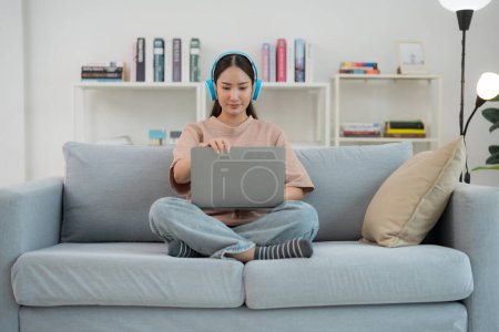 Mujer adulta joven contenta escucha música a través de auriculares mientras usa cómodamente su computadora portátil en un acogedor sofá, rodeado de un interior moderno con estanterías en el fondo