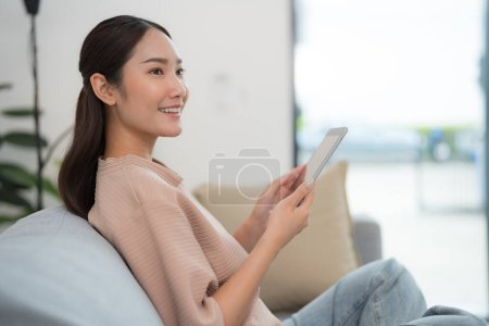 Foto de Hermosa joven asiática sentada cómodamente en un sofá, sonriendo a la cámara mientras usa una tableta digital en un ambiente de sala de estar moderno y bien iluminado - Imagen libre de derechos