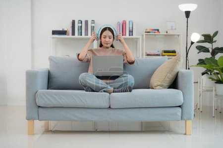 Zufriedene junge erwachsene Frau hört Musik über Kopfhörer, während sie bequem ihren Laptop auf einem gemütlichen Sofa bedient, umgeben von einem modernen Wohnbereich mit Bücherregalen im Hintergrund