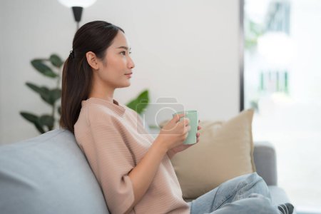 Foto de Mujer joven se sienta cómodamente en un sofá, sosteniendo una taza de café con una sonrisa suave, mirando hacia otro lado con un aire de satisfacción en una sala de estar moderna brillantemente iluminada - Imagen libre de derechos
