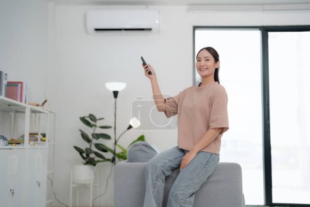 Young woman comfortably seated on a couch is using a remote control to adjust the air conditioner in a bright, modern living room with minimalist decor