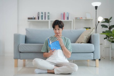 Focused young man sits cross-legged on the floor, reading a book with deep interest in a bright, contemporary living room, embodying a moment of quiet leisure and intellectual engagement