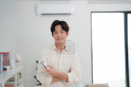 Junger Mann im lässigen Hemd mit einer Fernbedienung, um die Temperatur der Klimaanlage in einem hellen, modernen Wohnzimmer zu regulieren