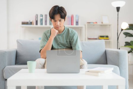 Besinnlicher junger Mann, der auf einer Couch sitzt, arbeitet an seinem Laptop in einem gut beleuchteten Wohnzimmer, wirkt konzentriert und leicht besorgt, mit einer Tasse und Büchern in der Nähe