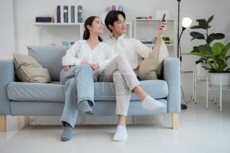 Glückliches junges Paar lümmelt auf einem Sofa in einem hellen, modernen Wohnzimmer, mit einer Fernbedienung, um die Klimaanlage für erhöhten Wohnkomfort anzupassen