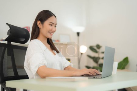 Die junge, lächelnde Frau sitzt gemütlich an ihrem Schreibtisch und arbeitet an ihrem Laptop in einem hellen, modernen Büroraum, der Professionalität und Konzentration ausstrahlt.