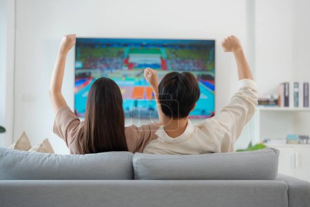 Back view of an enthusiastic couple sitting on a sofa, arms raised in victory while watching a live sports match on their home television, expressing excitement and support