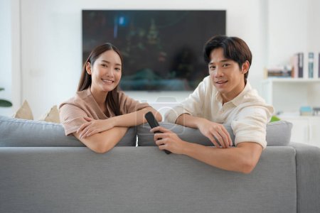 Junges Paar entspannt sich auf einem Sofa in einem hellen Wohnzimmer mit einem gedämpften Fernseher im Hintergrund und ruft Gefühle des Komforts, des Glücks und des lockeren Familienlebens hervor