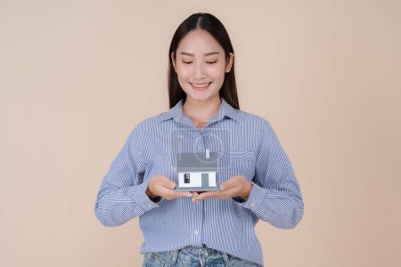 Photo for Cheerful young woman in a blue striped shirt confidently presents a small house model to the camera, symbolizing home ownership, real estate investment, or architectural concept - Royalty Free Image