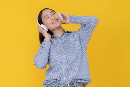 Foto de Mujer asiática joven enérgica en una camisa a rayas y jeans baila con auriculares, ojos cerrados, expresando felicidad y espíritu despreocupado contra un vibrante telón de fondo amarillo - Imagen libre de derechos
