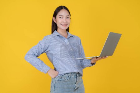 Foto de Mujer asiática joven elegantemente vestida con una camisa de rayas azules sonríe mientras sostiene un portátil gris, de pie sobre un vibrante telón de fondo amarillo, exudando confianza y profesionalismo - Imagen libre de derechos