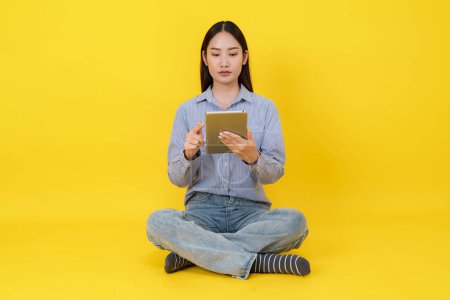 Fröhliche junge Asiatin sitzt im Schneidersitz auf leuchtend gelbem Hintergrund und lächelt, während sie mit einem digitalen Tablet hantiert, das Konnektivität und die Freude an der Technik in lässiger Kleidung symbolisiert