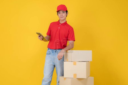 Cheerful delivery man in a red polo and cap stands beside a stack of packages, holding a digital tablet on a vibrant yellow background, depicting efficiency and online order management