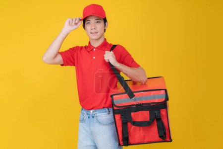 Porträt eines lächelnden jungen Auslieferers mit roter Mütze und Poloshirt, der eine isolierte Essensausgabetasche vor einem leuchtend gelben Hintergrund hält und damit einen effizienten Essenslieferservice symbolisiert