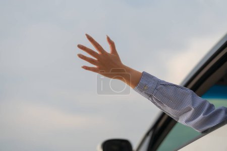Person's arm extends out of a car window waving against a clear sky, illustrating themes of travel, goodbyes, greetings, or seeking attention
