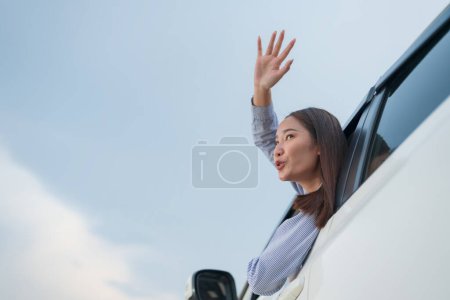 Foto de Mujer joven despreocupada agita su mano fuera de la ventana del coche, saludando al mundo con emoción contra un fondo de cielo suave durante una aventura de viaje por carretera - Imagen libre de derechos