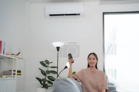 Jeune femme souriante utilisant son smartphone dans un salon bien éclairé pour contrôler à distance un climatiseur mural élégant, illustrant la technologie de la maison intelligente et la commodité moderne