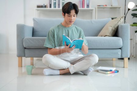 Focused young man sits cross-legged on the floor, reading a book with deep interest in a bright, contemporary living room, embodying a moment of quiet leisure and intellectual engagement