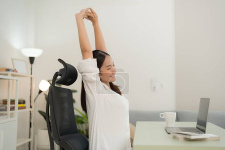 Foto de Mujer asiática en una blusa blanca toma un refrescante descanso de estiramiento en su configuración de oficina en el hogar minimalista, con una computadora portátil, taza de café y un fondo luminoso y aireado que mejora su postura relajada - Imagen libre de derechos