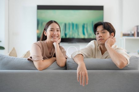 Young couple relaxes on a sofa in a bright living room with a muted television in the background, evoking feelings of comfort, happiness, and casual home life
