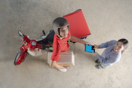 Draufsicht eines freundlichen Zustellers in roter Uniform bei der Übergabe eines Pakets an eine lächelnde Kundin, die auf einem mobilen Gerät unterschreibt, im Hintergrund ein Roller mit Zustellbox