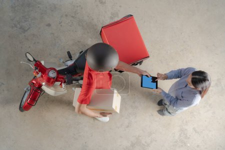 Draufsicht eines freundlichen Zustellers in roter Uniform bei der Übergabe eines Pakets an eine lächelnde Kundin, die auf einem mobilen Gerät unterschreibt, im Hintergrund ein Roller mit Zustellbox