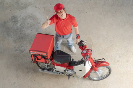 Top view of a cheerful delivery man in red uniform giving a thumb up next to his red motorcycle with an insulated box attached, ready for speedy food delivery service