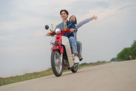 Young asian couple in casual clothing enjoying a fun ride on a red motorcycle on an open road, with woman raising arms feeling free and happy