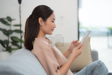 Foto de Hermosa joven asiática sentada cómodamente en un sofá, sonriendo a la cámara mientras usa una tableta digital en un ambiente de sala de estar moderno y bien iluminado - Imagen libre de derechos
