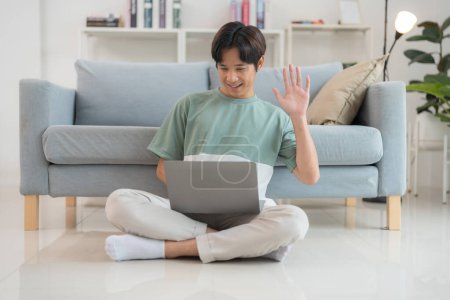 Foto de Joven hombre asiático se sienta con las piernas cruzadas en el suelo, se centró en su computadora portátil en una sala de estar moderna y luminosa, que simboliza el trabajo remoto, la comodidad y la tecnología en la vida cotidiana - Imagen libre de derechos