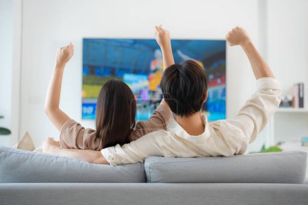 Back view of an enthusiastic couple sitting on a sofa, arms raised in victory while watching a live sports match on their home television, expressing excitement and support