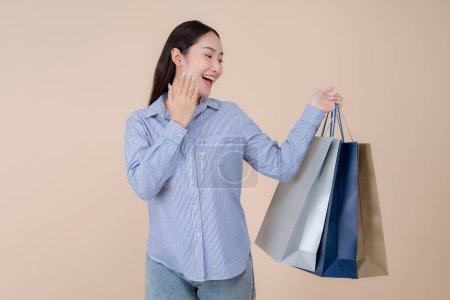 Foto de Joven mujer asiática alegre en ropa vaquera casual está sosteniendo múltiples bolsas de compras con una sonrisa radiante sobre un fondo pastel, expresando emoción y satisfacción después de ir de compras - Imagen libre de derechos