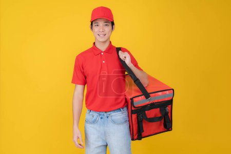Portrait of a smiling young delivery man wearing a red cap and polo shirt, holding an insulated food delivery bag against a vibrant yellow background, symbolizing efficient food delivery service