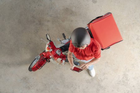 Vista superior de un mensajero de reparto en uniforme rojo y casco, de pie junto a un scooter rojo con una gran mochila aislada, mirando hacia arriba, presumiblemente esperando el siguiente pedido