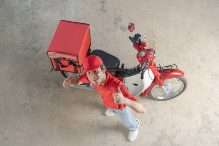 Top view of a cheerful delivery man in red uniform giving a thumb up next to his red motorcycle with an insulated box attached, ready for speedy food delivery service