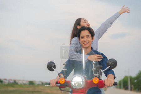 Foto de Joven pareja asiática en ropa casual disfrutando de un divertido paseo en una motocicleta roja en un camino abierto, con la mujer levantando los brazos sintiéndose libre y feliz - Imagen libre de derechos
