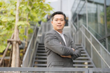 Foto de Profesional asiático hombre de negocios en un traje gris se levanta con los brazos cruzados al aire libre, exudando confianza con un edificio de oficinas urbano escalera fondo y vegetación - Imagen libre de derechos