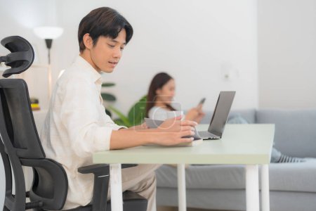 Hombre joven centrado en el trabajo en un escritorio minimalista con ordenador portátil en una oficina en casa brillante, mientras que una mujer se relaja en un sofá en el fondo, encarnando el equilibrio entre el trabajo y la vida