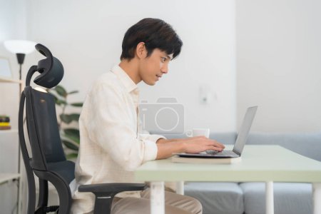 Foto de Vista lateral de un joven enfocado escribiendo en una computadora portátil en un entorno de oficina en casa brillante y moderno, con una taza de café y una silla cómoda, ejemplificando el trabajo remoto - Imagen libre de derechos