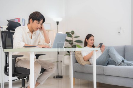 Young man focused on work at a minimalist desk with laptop in a bright home office, while a woman relaxes on a couch in the background, embodying work-life balance