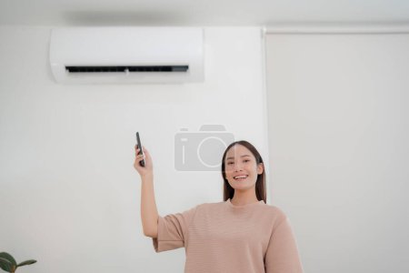 Foto de Mujer joven sonriente en una camisa casual de pie en una sala de estar luminosa, ajustando con confianza la temperatura del aire acondicionado con un control remoto, disfrutando de un estilo de vida cómodo y moderno en el interior - Imagen libre de derechos