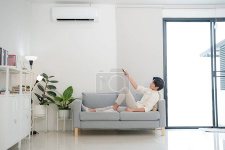 Young man sits on a sofa, comfortably controlling the air conditioning with a remote, in a well-lit and modern living room, showcasing a lifestyle of convenience and technology