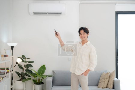 Smiling young man in a casual shirt standing in a bright living room, confidently adjusting the air conditioner temperature with a remote control, enjoying a comfortable and modern lifestyle indoors