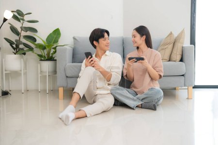 Young, smiling couple sits comfortably on a sofa, each engrossed in their own smartphone, sharing a moment of digital leisure in a bright, cozy living room setting