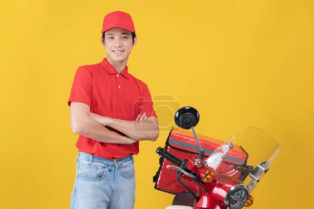 Cheerful young delivery man stands casually beside a red motorcycle. Arms crossed. Wearing a red uniform with a cap. Set against a vibrant yellow backdrop